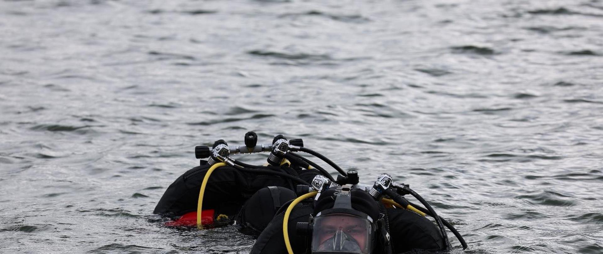 Na zdjęciu widać dwóch nurków w wodzie. Obaj mają na sobie czarne pianki, maski z pełnym zakryciem twarzy oraz sprzęt do nurkowania z butlami tlenowymi i żółtymi wężami. Woda jest spokojna, a nurkowie unoszą się na jej powierzchni, częściowo zanurzeni. Tło stanowi rozległa tafla wody bez widocznego brzegu.