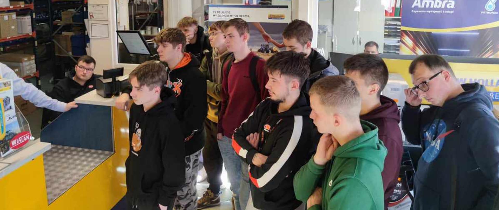 Uczniowie stoją przy ladzie w dziale „Części i Akcesoria” firmy Wialan, rozmawiając z pracownikiem siedzącym przy komputerze.