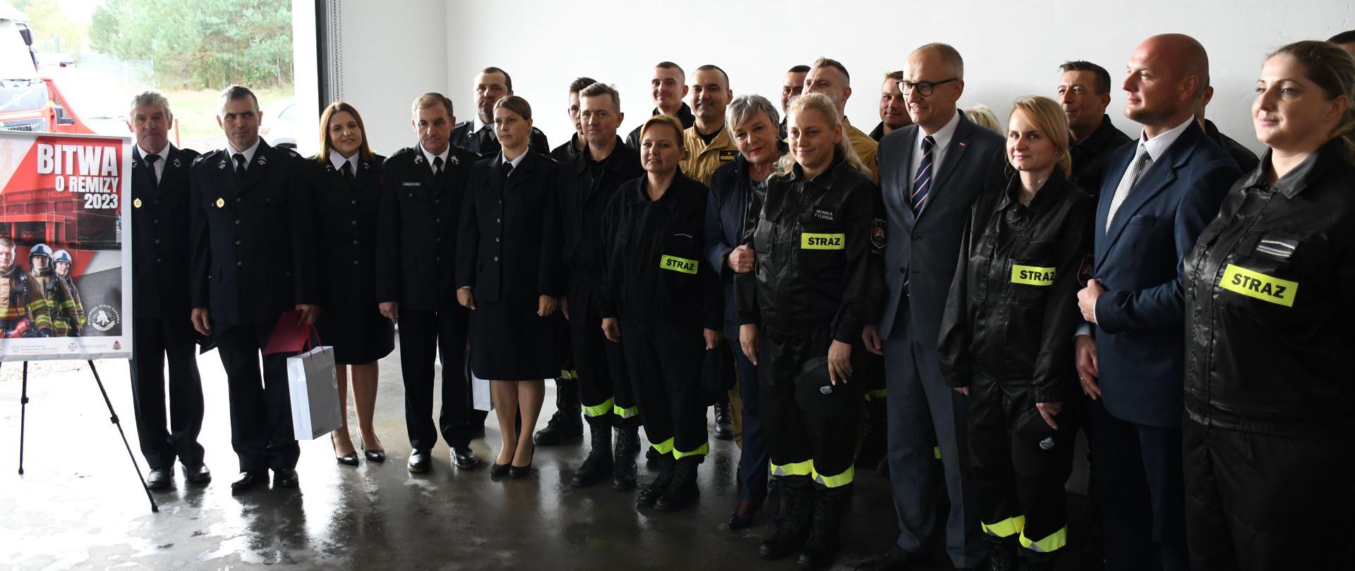 Zdjęcie przedstawia uczestników uroczystości wręczenia promes na sfinansowanie zakupu sprzętu pożarniczego jednostkom OSP w Surminach i Baniach Mazurskich, ktorzy są zlokalizowani w garażu Ochotniczej Straży Pożarnej w Surminach
