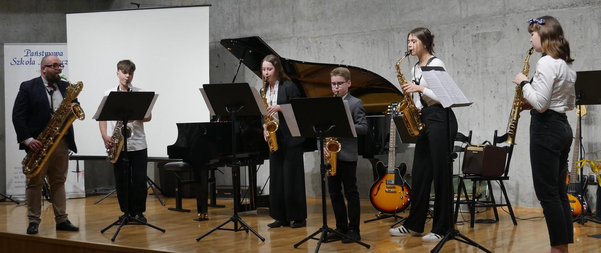 Grupa uczniów i nauczyciel grają na scenie na saksofonach