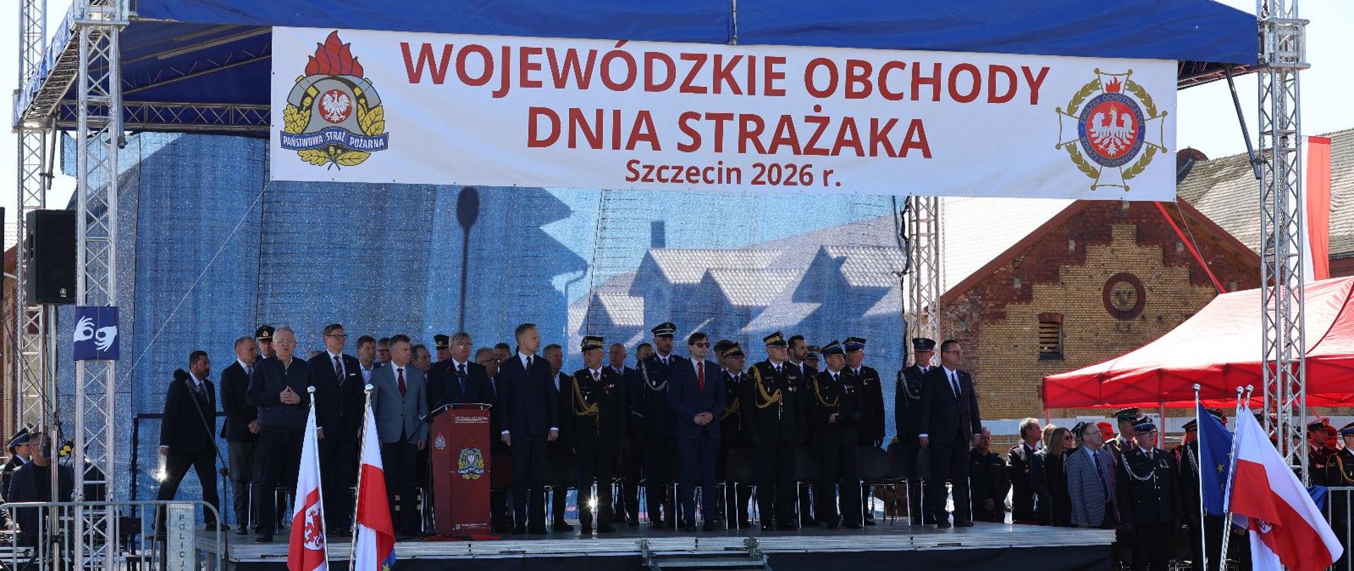 Scena podczas Wojewódzkich Obchodów Dnia Strażaka w Szczecinie. Na podwyższeniu pod niebieskim zadaszeniem stoją zaproszeni goście, funkcjonariusze Państwowej Straży Pożarnej oraz poczty sztandarowe. Nad sceną widoczny jest baner z napisem „Wojewódzkie Obchody Dnia Strażaka Szczecin 2026 r.”.