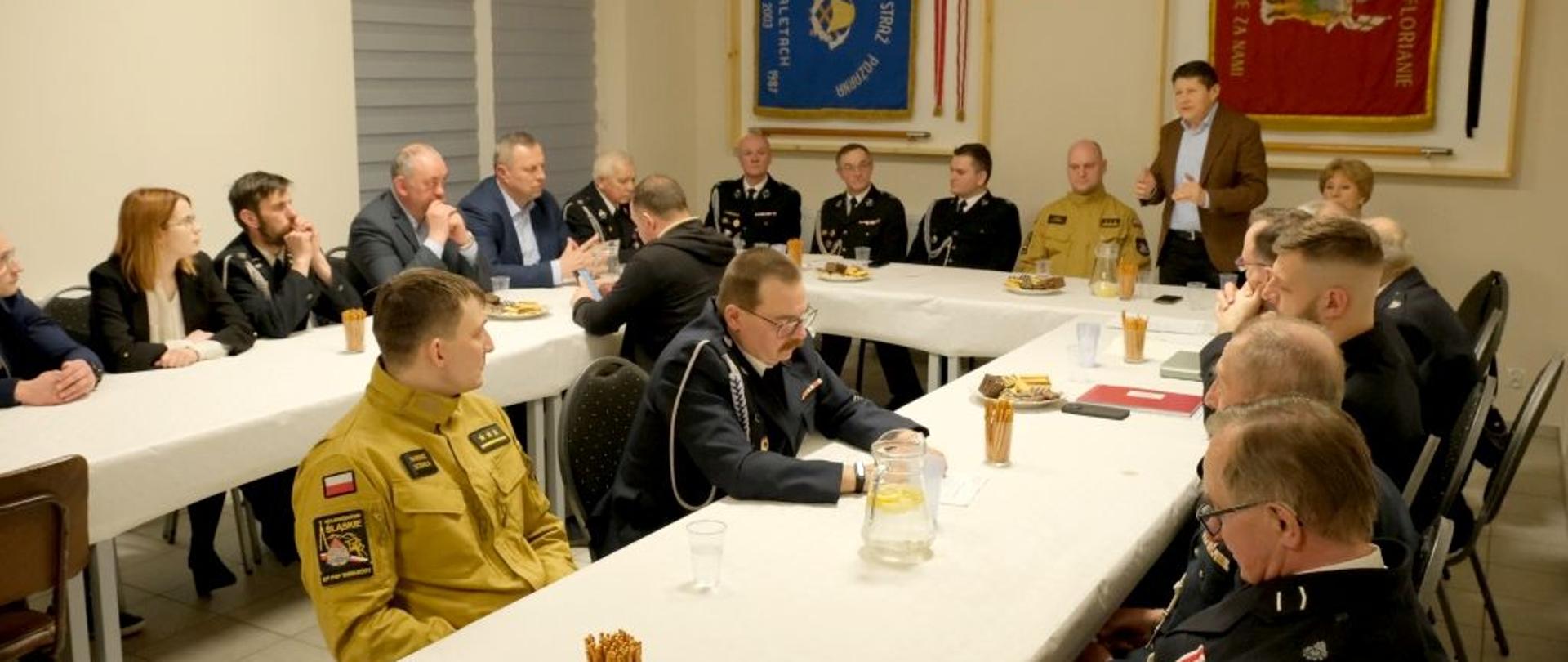 Na sali przy stole w kształcie litery U siedzą strażacy w mundurach i osoby ubrane po cywilnemu. Jedna osoba stoi i przemawia.