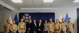 Grupa strażaków PSP w żółtych mundurach służbowych oraz osób cywilnych pozuje wspólnie do zdjęcia grupowego w sali konferencyjnej Komendy Głównej PSP. W tle widoczne są flagi Polski i Unii Europejskiej oraz logo PSP.