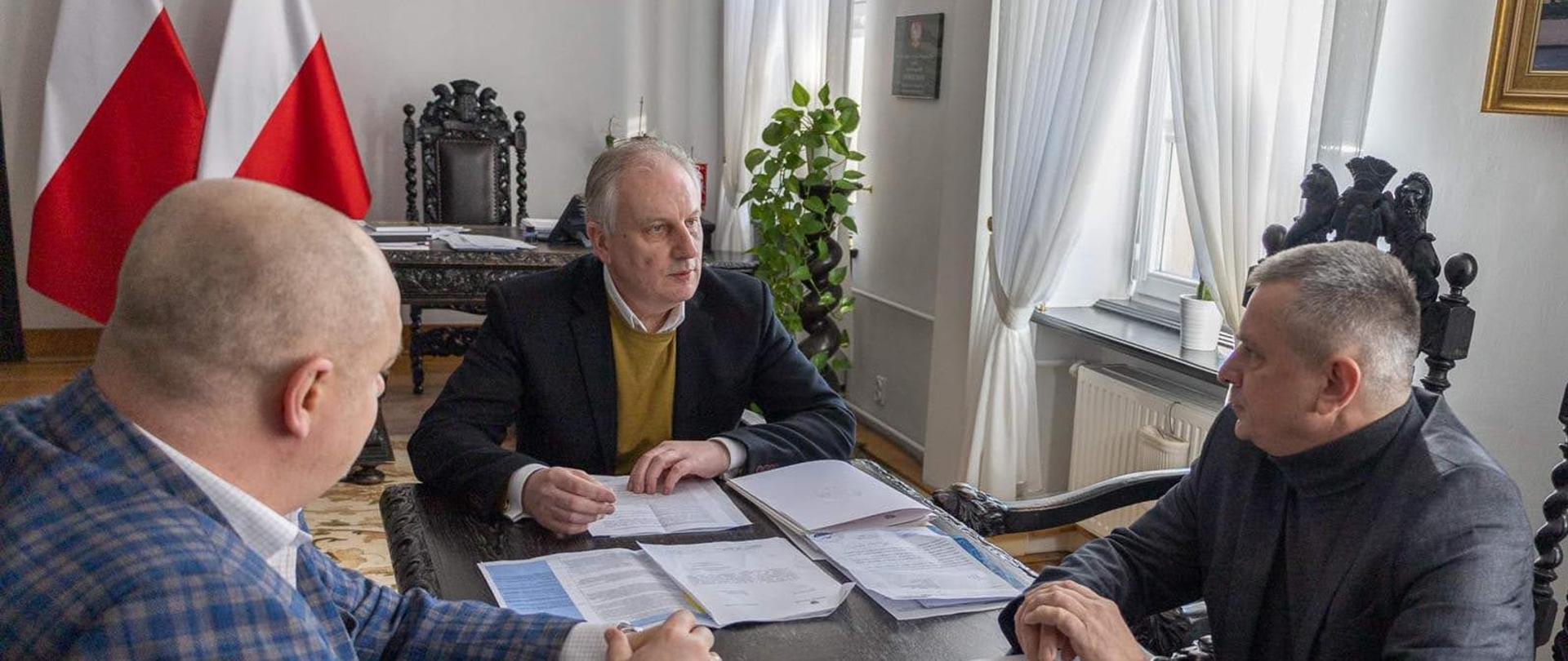 Wojewoda pomorski, pomorski komendant wojewódzki Państwowej Straży Pożarnej oraz komendant wojewódzki Policji w Gdańsku rozmawiają przy stole na którym znajdują się dokumenty. 