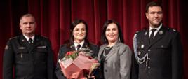 Podczas uroczystości Mundurowego Dnia Kobiet uhonorowana starszy aspirant Joanna Jatczak- Kuczkowska pozuje wspólnie do zdjęcia z Podsekretarzem Stanu Adamem Nowakiem, Wicemarszałek Województwa Łódzkiego Agnieszkom Ryś i Zastępom Łódzkiego Komendanta Wojewódzkiego Państwowej Straży Pożarnej starszym brygadierem Wojciechem Wielochem. Starszy aspirant Joanna Jatczak- Kuczkowska w ręku trzyma bukiet kwiatów. 