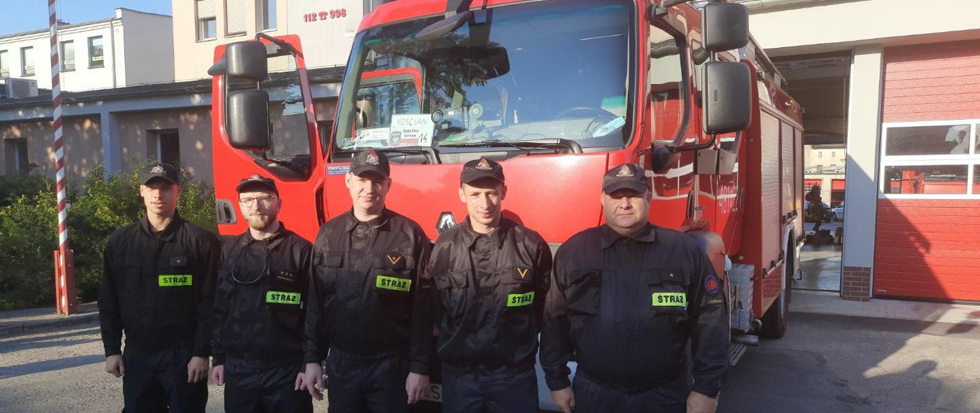 Strażacy z Polski pomogą w walce z pożarami w Grecji. Strażacy, którzy wyjadą do Grecji stoją przed samochodem pożarniczym. w tle budynek komendy. 