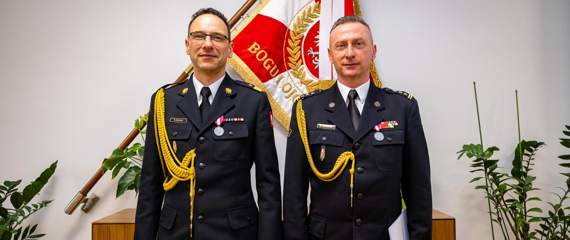 Oficerowie PSP stoją obok siebie w mundurach galowych, prezentując odznaczenia i dystynkcje służbowe. Za nimi widoczny jest sztandar PSP oraz elegancka aranżacja biurowa. Zdjęcie podkreśla oficjalny charakter spotkania, prawdopodobnie związanego z awansem, odznaczeniem lub zmianą na stanowisku dowódczym.