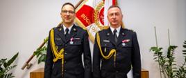 Oficerowie PSP stoją obok siebie w mundurach galowych, prezentując odznaczenia i dystynkcje służbowe. Za nimi widoczny jest sztandar PSP oraz elegancka aranżacja biurowa. Zdjęcie podkreśla oficjalny charakter spotkania, prawdopodobnie związanego z awansem, odznaczeniem lub zmianą na stanowisku dowódczym.