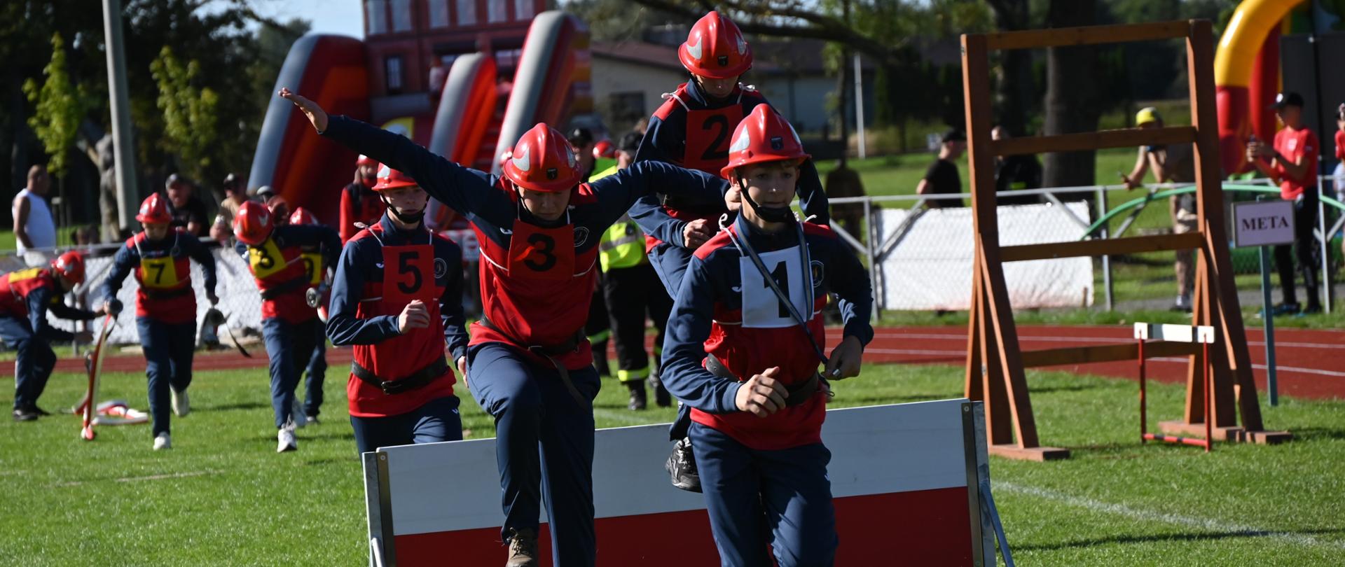 Grupa młodych strażaków w dresach i hełmach czerwonych przeskakuje przez ściankę. W tle inne chłopaki rozwijają linię gaśniczą.