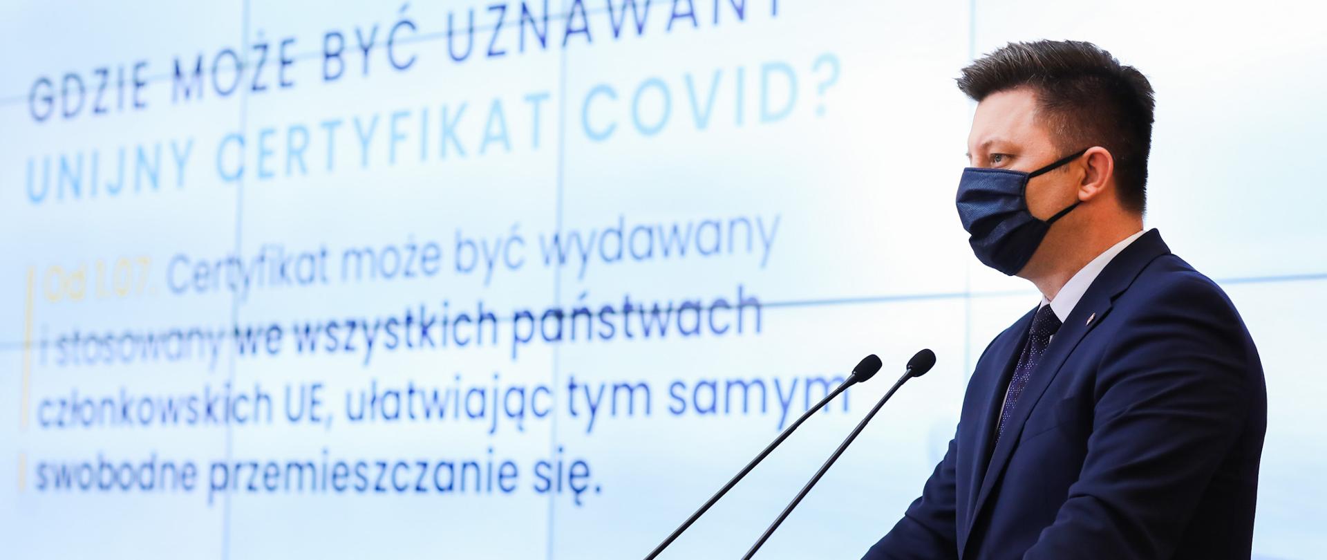 Szef KPRM Michał Dworczyk stoi za pulpitem z mikrofonami podczas konferncji dot. szczepień dzieci i unijnych certyfikatów Covid.