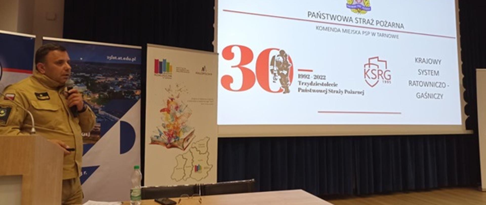 Na zdjęciu widoczny jest funkcjonariusz PSP podczas prelekcji w Akademii Nauk Stosowanych. Funkcjonariusz omawia funkcjonowanie Państwowej Straży Pożarnej, a w ręce trzyma mikrofon. Na ekranie wyświetlony jest slajd informujący o trzydziestej rocznicy powstania Państwowej Straży Pożarnej. 
