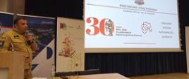 Na zdjęciu widoczny jest funkcjonariusz PSP podczas prelekcji w Akademii Nauk Stosowanych. Funkcjonariusz omawia funkcjonowanie Państwowej Straży Pożarnej, a w ręce trzyma mikrofon. Na ekranie wyświetlony jest slajd informujący o trzydziestej rocznicy powstania Państwowej Straży Pożarnej. 