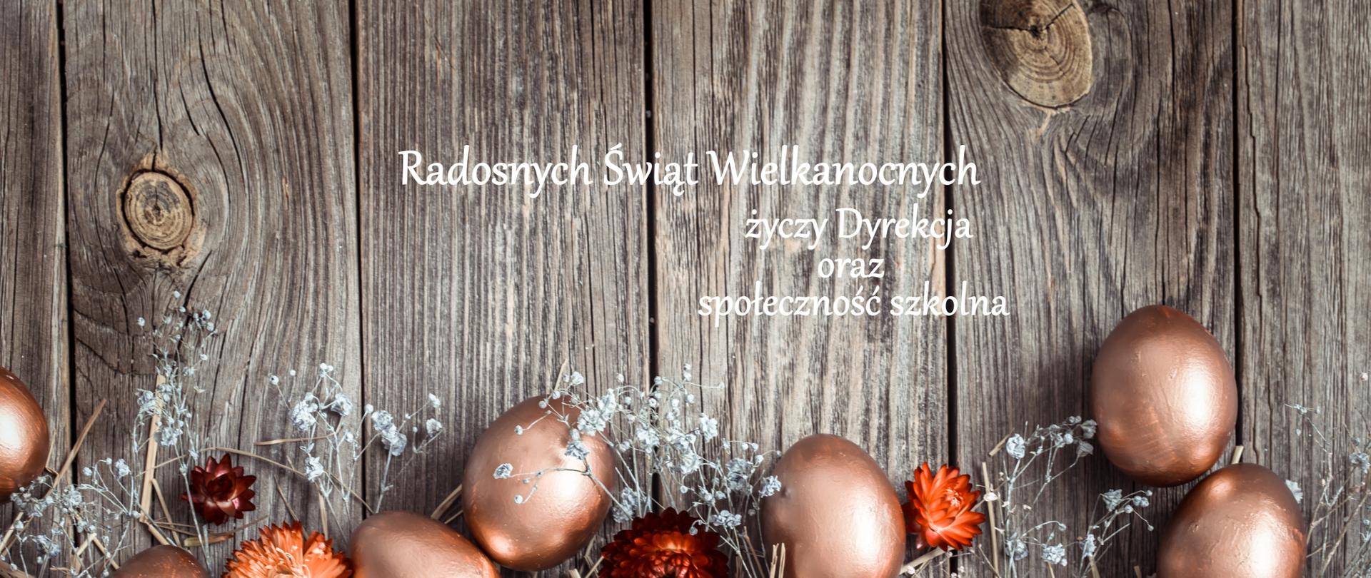Na obrazie widać dekoracyjną, świąteczną kompozycję utrzymaną w ciepłych, naturalnych kolorach.
Tło stanowi pionowa, drewniana ściana z wyraźną fakturą słojów i sęków, w odcieniach szaro-brązowych. Na dole, na warstwie jasnej słomy, ułożone są liczne jajka pomalowane na metaliczny, miedziano-złoty kolor. Mają gładką, lekko błyszczącą powierzchnię i są rozmieszczone nieregularnie, tworząc dekoracyjny pas wzdłuż dolnej części obrazu.
Między jajkami znajdują się suszone kwiaty w delikatnych kolorach: kremowym, jasnoróżowym oraz intensywnie pomarańczowym i czerwonym. Widać też cienkie, białe gałązki przypominające drobne, suche rośliny ozdobne.
W górnej części obrazu, na drewnianym tle, umieszczony jest biały napis w języku polskim:
„Radosnych Świąt Wielkanocnych
życzy Dyrekcja
oraz społeczność szkolna”.
Całość ma spokojny, elegancki i lekko rustykalny charakter, kojarzący się z Wielkanocą i wiosenną dekoracją.