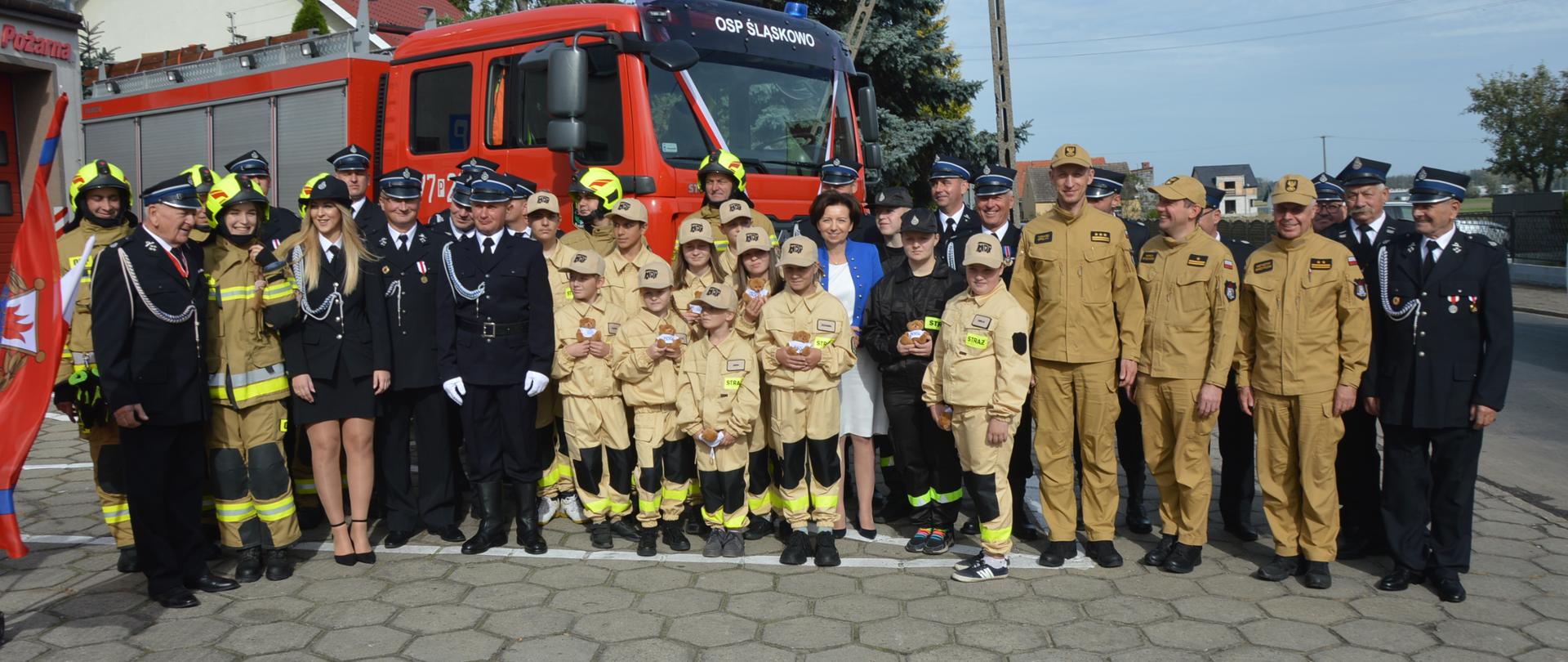 Przekazanie pojazdu ratowniczo-gaśniczego do OSP Śląskowo. Zdjęcie przedstawia plac przed remizą OSP w Śląskowie. Na tle przekazanego pojazdu, do wspólnego zdjęcia pozują uczestnicy uroczystości: pani minister, strażacy OSP i PSP oraz członkowie młodzieżowej drużyny pożarniczej. 