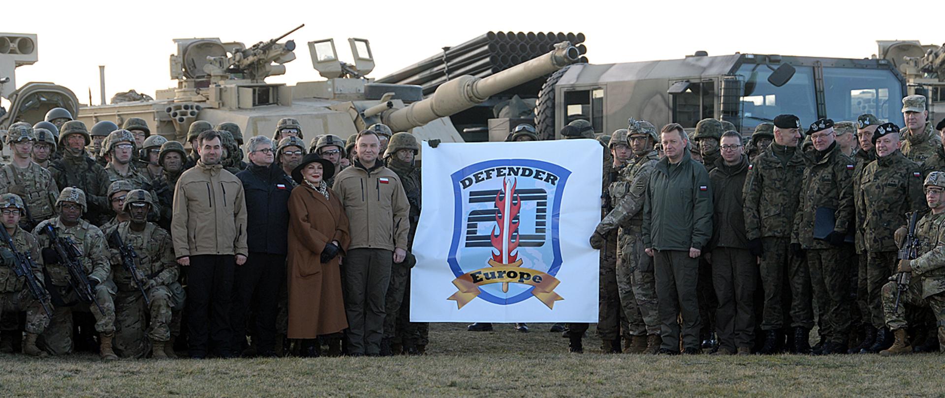 Zdjęcie grupowe prezydenta i ministra obrony narodowej z żołnierzami polskimi i amerykańskimi biorącymi udział w ćwiczeniu Defender Europe 20 w Drawsku Pomorskim. w tle sprzęt wojskowy.