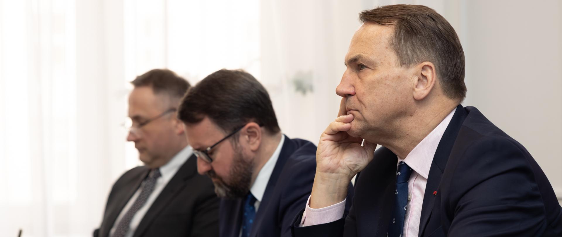 Na zdjęciu jest trzech mężczyzn w garniturach. Siedzą przy stole. Robią notatki. 