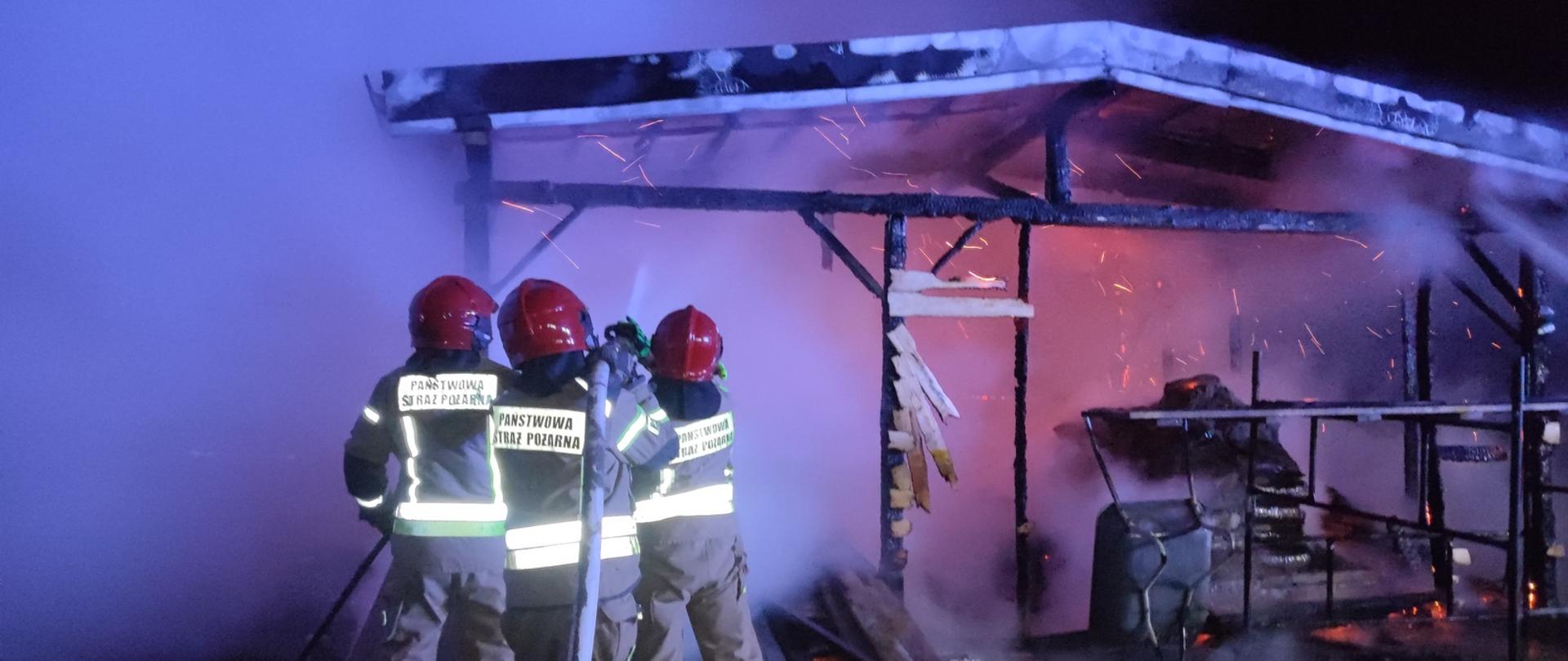 Zdjęcie wykonane w porze nocnej na miejscu pożaru szopy. Trzech strażaków ubranych w piaskowe ubranie specjalne z napisem Państwowa Straż Pożarna oraz czerwone hełmy gaszą szopę. W tle widać gęsty biały dym oraz iskry wydobywające się z szopy.