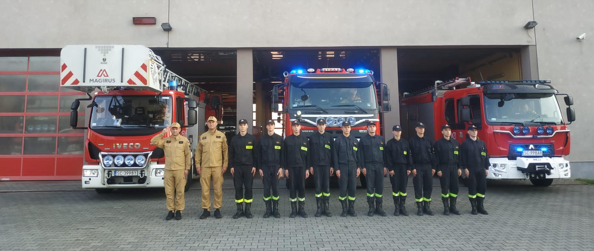 Strażacy ze zmiany JRG ustawieni w szeregu przed bramami garażowymi jednostki, za nimi widoczne samochody bojowe z włączonymi sygnałami świetlnymi