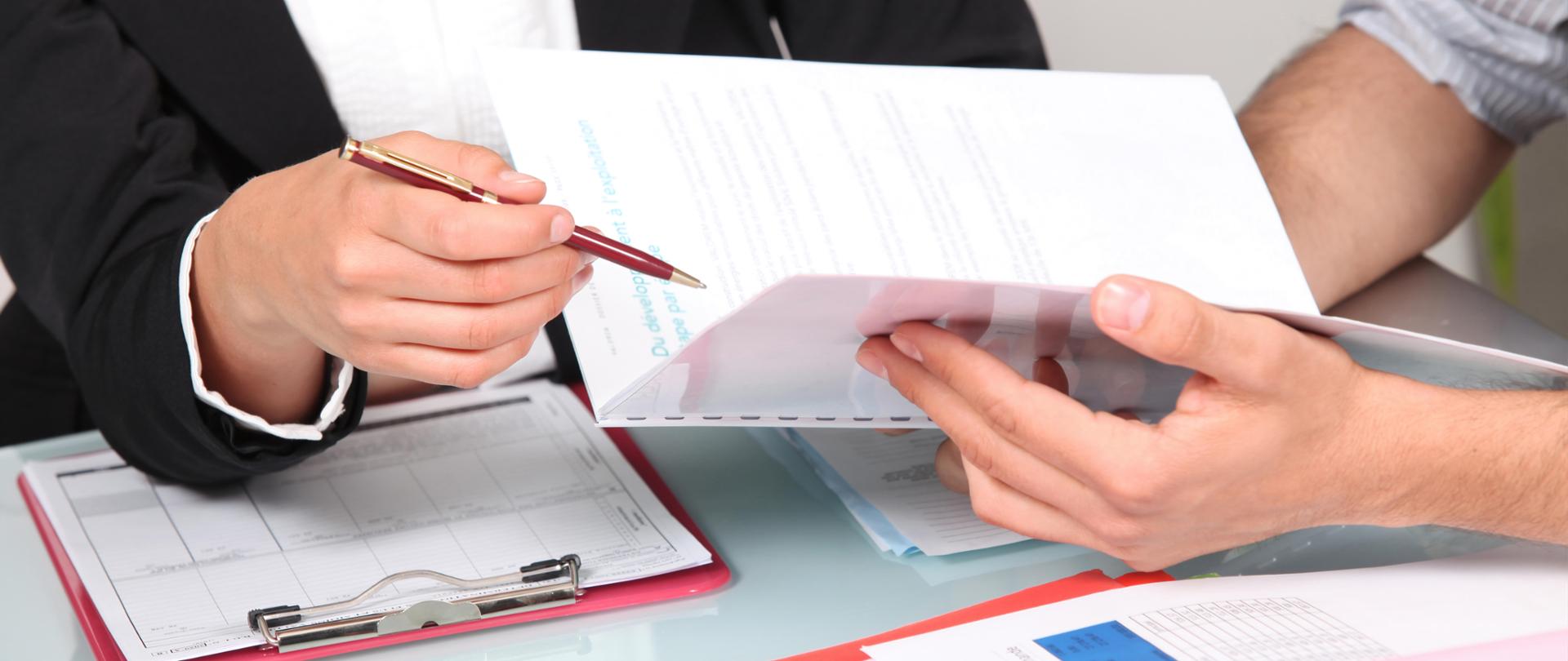 Mężczyzna nad biurkiem trzyma w dłoni zbindowane kartki papieru na które wskazuje kobieta długopisem