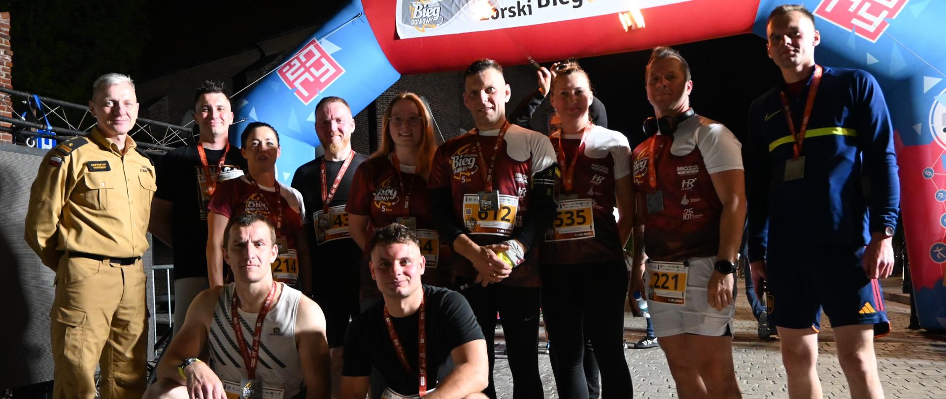 grupa uczestników XIII Biegu Ogniowego pozuje do wspólnego zdjęcia przy bramie wydarzenia.