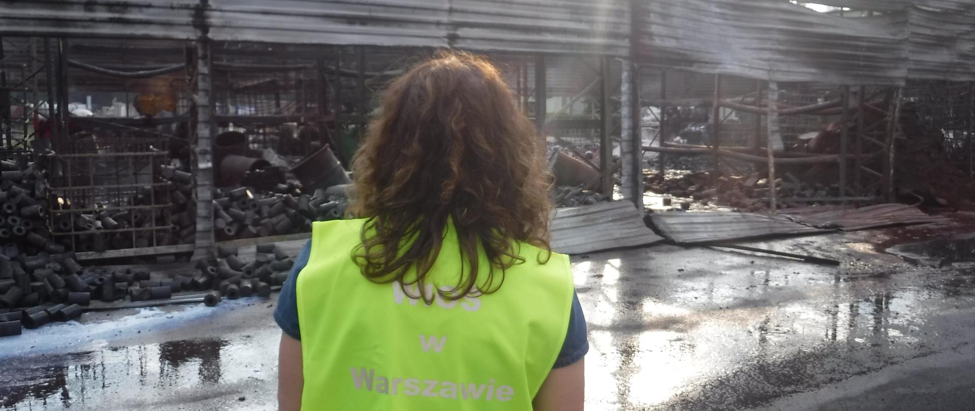 Inspektorka Wojewódzkiego Inspektoratu Ochrony Środowiska w Warszawie prowadzi działania na terenie pożaru. Obserwuje wysoką na kilka metrów szarą, stalową halę stojącą na zalanej wodą wylewce betonowej i znajdującą się w odległości kilku metrów od inspektorki. Dolna cześć hali pozbawiona jest ściany i wewnątrz widać spalone pojemniki.