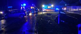 Zdjęcie przedstawia dwa rozbite samochody osobowe, jeden w przydrożnym rowie , drugi na jezdni. W tle widać dwie karetki pogotowia, strażaków i wóz strażacki stojące z włączonymi niebieskimi światłami 