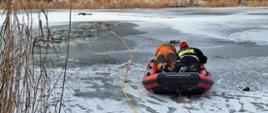 Na zdjęciu pokrywa lodowa na jeziorze. W tle trzciny. Na pierwszym planie specjalistyczna deska, przypominająca nadmuchiwany ponton, na której „na brzuchu” leżą dwie osoby spoglądające w dal przed siebie. Po lewej w pomarańczowo-czarnym kombinezonie, po prawej w czerwono-czarnej czapce na głowie, w czarnym ubraniu z naszytym na plecach żółtym prostokątem, na którym widać fragment napisu: Straż Pożarna. Do czerwono-czarnej deski przyczepiona czerwona lina. Druga żółta lina biegnie po lodzie w głąb zbiornika wodnego. W oddali ledwie widoczna czarna postać leżąca na wznak na lodzie.