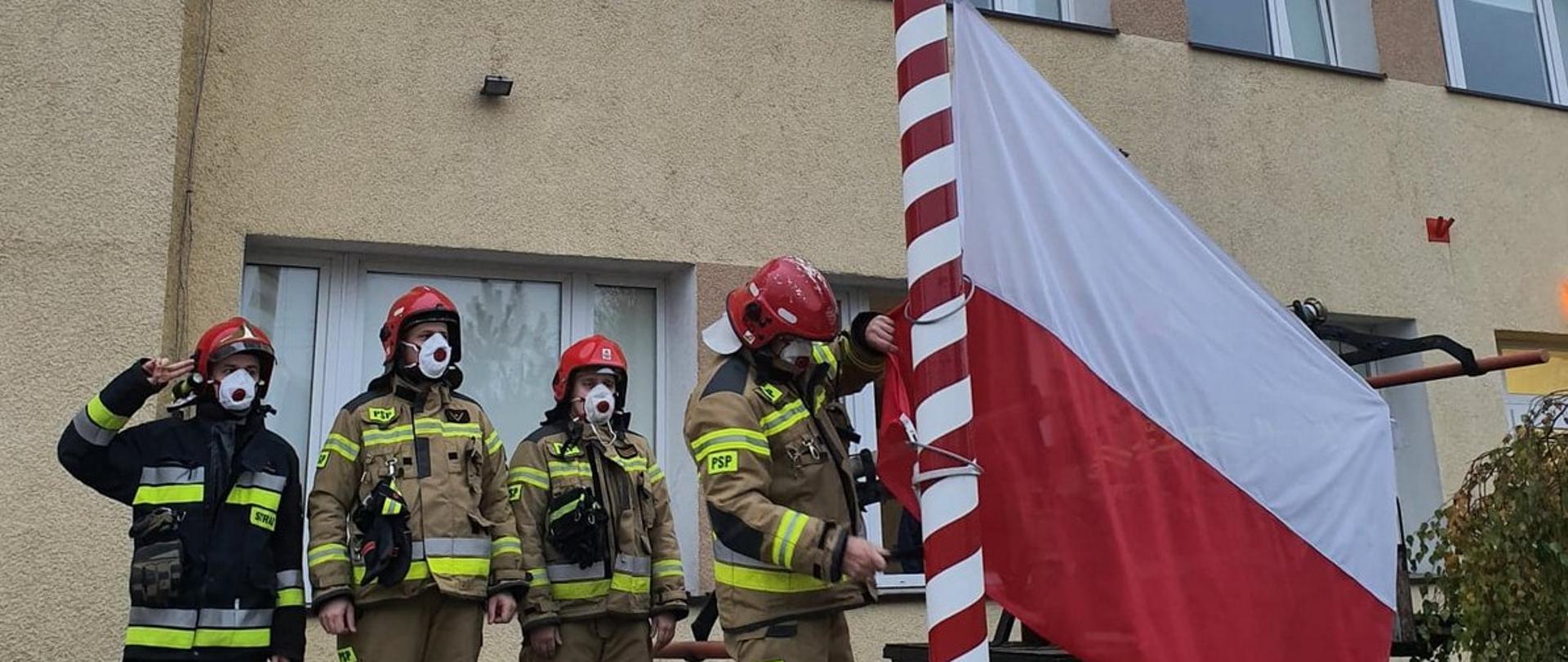 Zdjęcie zrobione na tle budynku komendy, przed masztem flagowym z flagą na dole przed wciągnięciem na maszt. Strażacy w ubraniach specjalnych w hełmach stoją w szeregu na baczność.