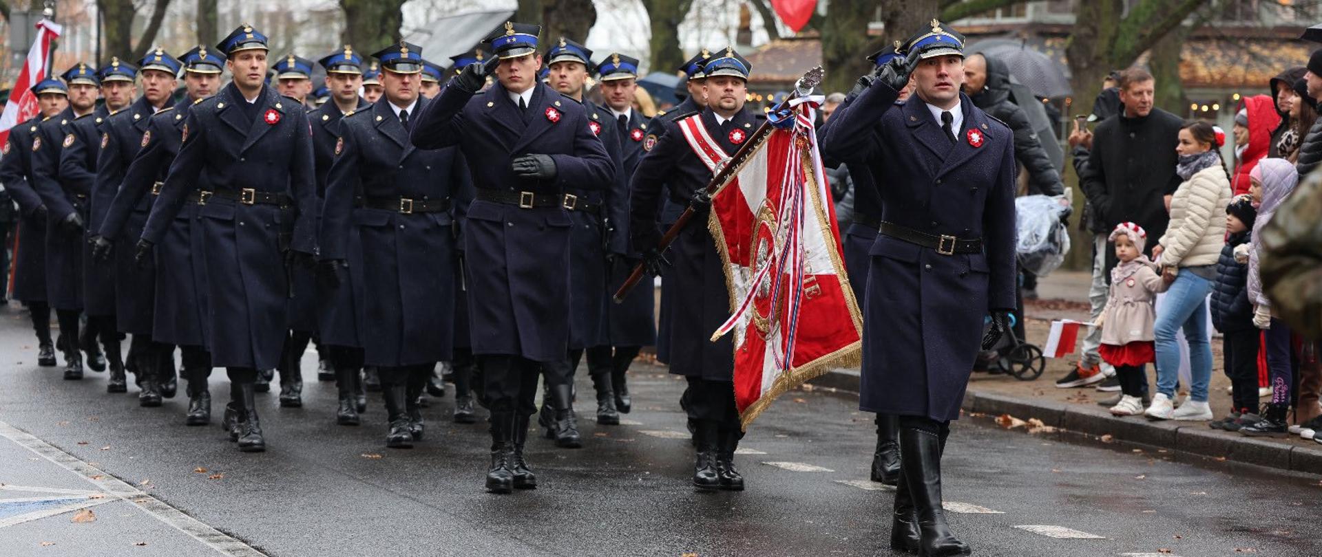 Na zdjęciu widać uroczystą paradę odbywającą się na ulicy w jesiennym otoczeniu. Strażacy ubrani w ciemnogranatowe płaszcze i czapki z daszkiem maszerują w równych szeregach. Na czele pochodu idzie oficer niosący sztandar ozdobiony biało-czerwonymi wstążkami. Kilku strażaków salutuje. W tle widać polskie flagi oraz grupę widzów — dorosłych i dzieci — obserwujących przemarsz z chodnika. Drzewa są bez liści, nawierzchnia jest mokra.