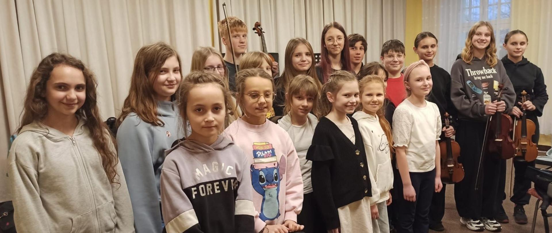 Zdjęcie kolorowe. Grupa dzieci i młodzieży stojąca w pomieszczeniu na tle jasnych zasłon. Uczestnicy ubrani w codzienne stroje: bluzy z kapturem, swetry i koszulki. Niektóre osoby trzymają instrumenty muzyczne – skrzypce oraz smyczki. W przednim rzędzie stoi dziewczynka w różowej bluzie z nadrukiem postaci z bajki, trzymająca czarny futerał na instrument. Po jej lewej stronie inna dziewczynka w szarej bluzie z napisem "MAGIC FOREVER". Osoby uśmiechają się lub patrzą bezpośrednio w stronę obiektywu. Po prawej stronie widoczne wysokie okno z firanką.