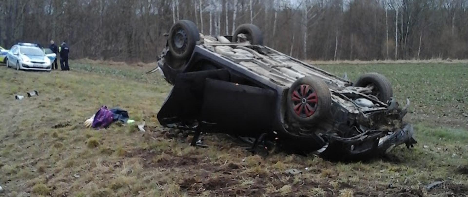 Zdjęcie przedstawia czarny samochód osobowy leżący na dachu, w tle srebrny samochód policyjny 