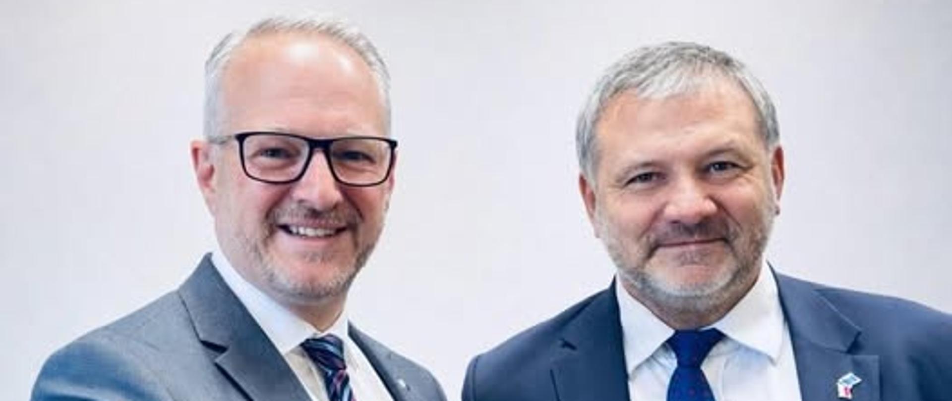 Dwóch mężczyzn, wojewoda i minister, podają sobie rękę, są ubrani w garnitury, uśmiechają się 