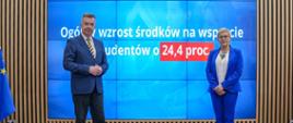 Minister Wieczorek i wiceminister Mrówczyńska stoją na sali, za nimi na wbudowanym w ścianę wielkim ekranie napis Ogólny wzrost środków na wsparcie studentów o 24,4 proc.