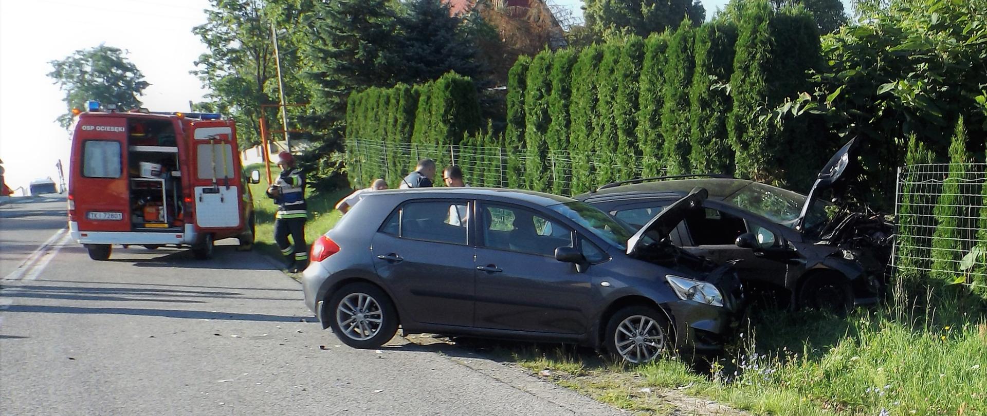 Zdjęcie przedstawia dwa samochody osobowe, które uległy zderzeniu niszcząc jednocześnie fragment ogrodzenia prywatnej posesji. W wyniku czego oba stoją na poboczu a za nimi widać ratowników i samochód pożarniczy biorący udział w likwidacji skutków wypadku.