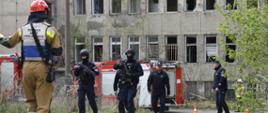 Policjanci w długą bronią w kominiarkach idący w stronę strażaka ustawionego tyłem do zdjęcia, w tle pojazdy pożarnicze