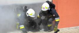 Dwóch strażaków w sprzęcie ochrony dróg oddechowych ewakuuje z zadymionego obiektu manekina.