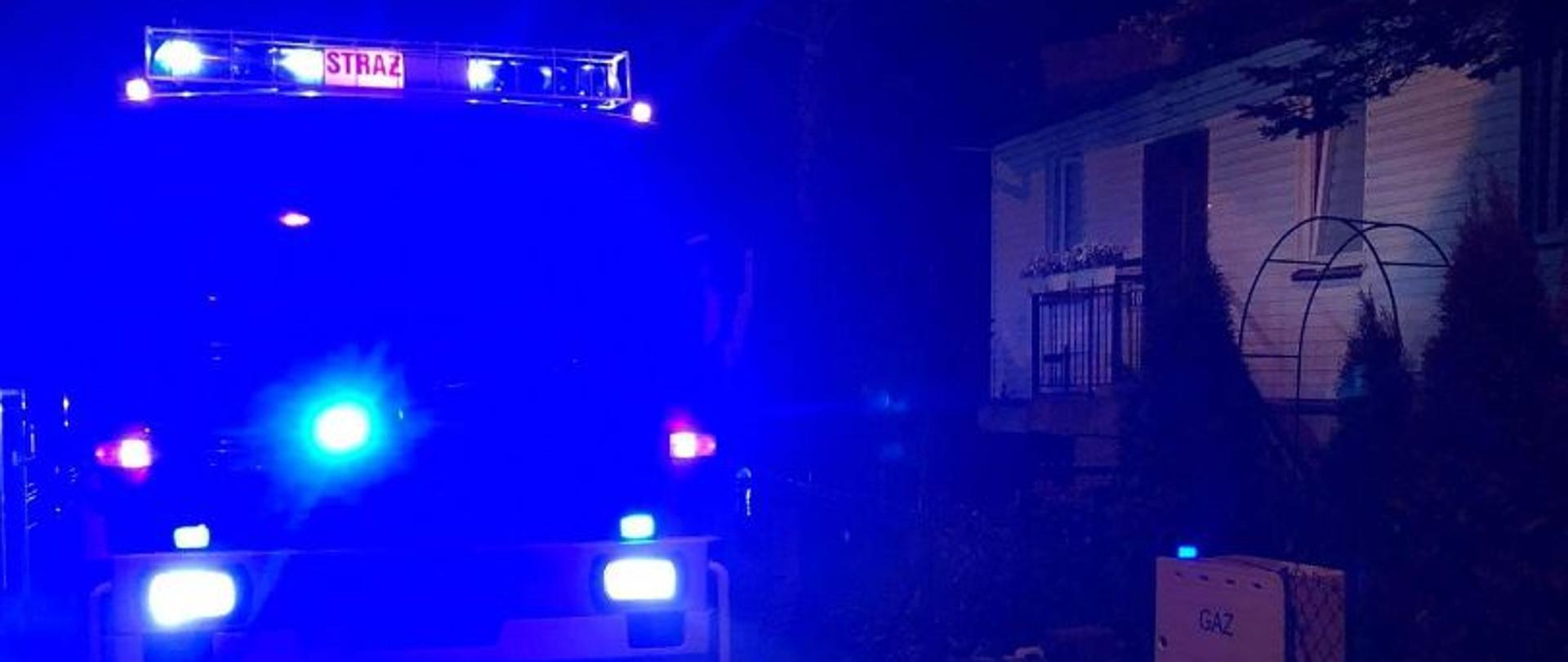 pojazd pożarniczy zaparkowany na ulicy przy budynku objętym pożarem w porze nocnej z zapalonymi niebieskimi światłami alarmowymi