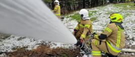 Kolorowa fotografia wykonana w pochmurny i śnieżny dzień na zewnątrz. Przedstawia strażaków OSP ubranych w ubrania specjalne i hełmy podających prąd gaśniczy z działka wodno – pianowego i prądownicy. W tle linie wężowe.