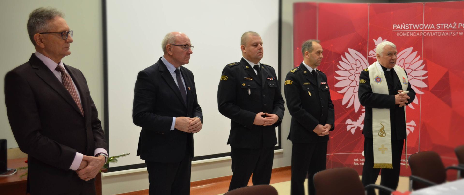 Na zdjęciu widoczni są Zachodniopomorski Komendant Wojewódzki PSP, Starosta Wałecki i Komendant Powiatowy PSP w Wałczu. Obok nich stoi Zachodniopomorski Kapelan, który odprawia modlitwę. Uroczystość odbywa się w sali konferencyjnej Komendy Powiatowej PSP. 