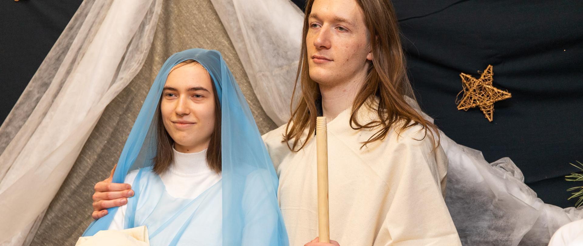 Zdjęcie przedstawia Józefa i Maryję z dzieciątkiem Jezus. Głowa Maryi przeplatana jest niebieskim tiulem, a Józef stoi po prawej stronie w lnianym kremowym ubraniu. Mężczyzna trzyma w ręku coś, co przypomina drewniany kij. Na ciemnym tle widać dekoracje ze złotych gwiazdek. 