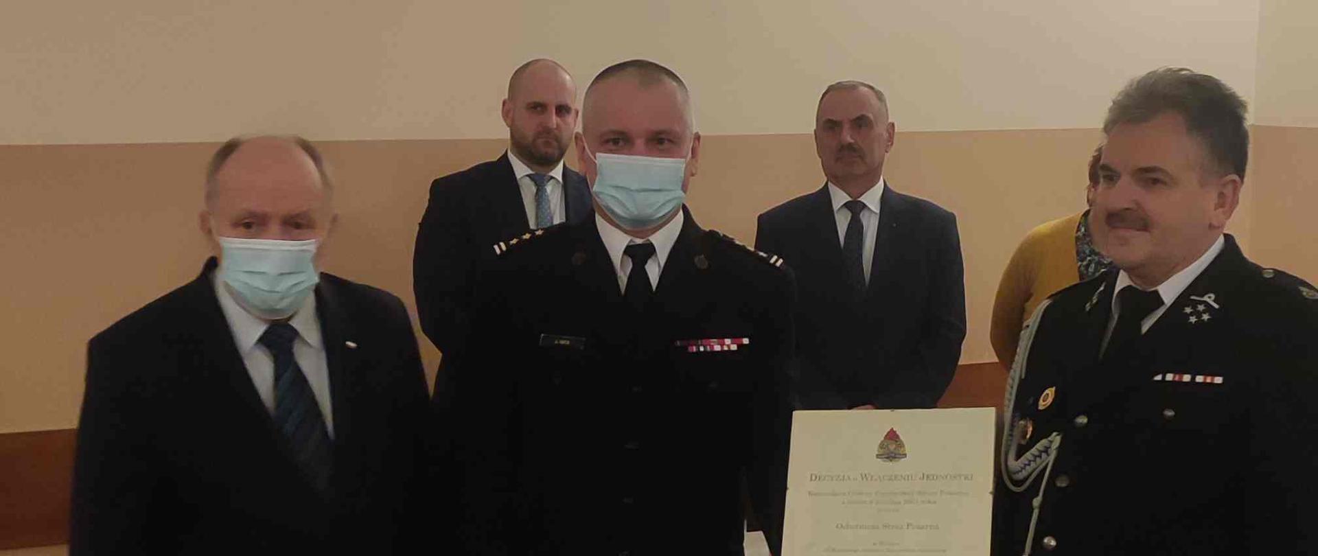 W pomieszczeniu znajduje się kilka osób. Na pierwszym planie stoi dwóch strażaków trzymających wspólnie dokument, obok nich osoba cywilna w garniturze. Za nimi dwóch mężczyzn w garniturach.