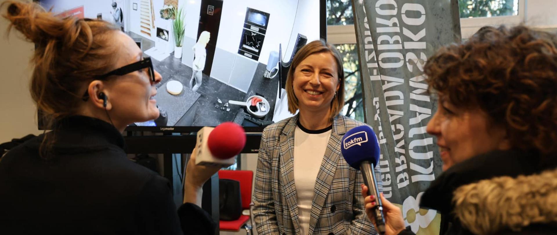 Kobieta, a przed nią dwie inne z mikrofonami. W tle baner reklamowy i duży ekran. 