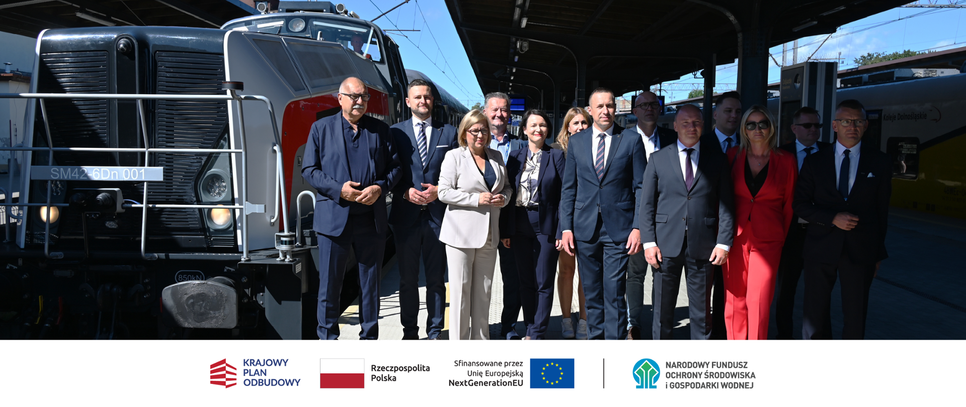 Zdjęcie na dworcu w Karpaczu - briefing prasowy. Widoczne są logotypy: KPO, Rzeczpospolita Polska (flaga), Unia Europejska – flaga UE z napisem „Sfinansowane przez Unię Europejską NextGenerationEU, Narodowy Fundusz Ochrony Środowiska i Gospodarki Wodnej.