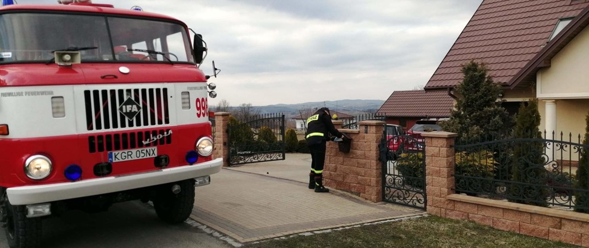 
Na zdjęciu widoczny druh Ochotniczej Straży Pożarnej wrzucający ulotkę „Szczepimy się” do skrzynki na listy zamontowanej w ogrodzeniu przed budynkiem jednorodzinnym. Po lewej jego stronie samochód strażacki IFA koloru czerwonego stojący na drodze asfaltowej. Na wprost budynek mieszkalny elewacja koloru pisakowego dach pokryty blachodachówką koloru brązowego.
