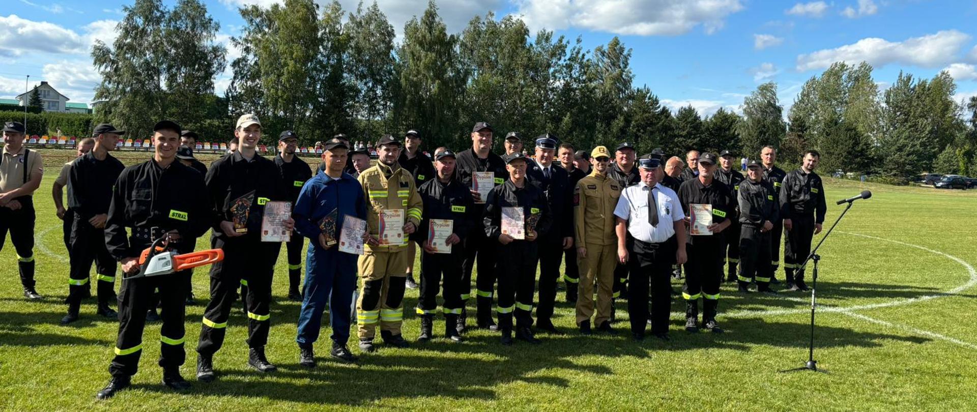 Zdjęcie przedstawia 11 mężczyzn, druhów OSP, stojących w rzędzie. Wszyscy ubrani w stroje koszarowe. Niektórzy z nich trzymają dyplomy oraz nagrody otrzymane podczas zawodów. Za nimi stoją pozostali członkowie drużyn. 