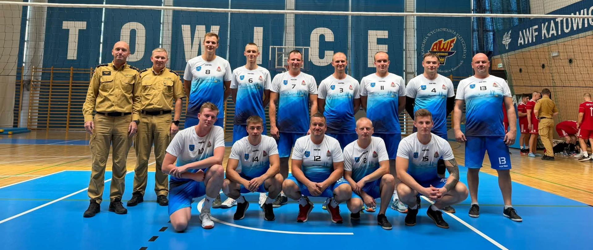 Na zdjęciu drużyna strażaków uczestnicząca w zawodach sportowych w piłce siatkowej. Zawodnicy w strojach sportowych pozują do wspólnego zdjęcia na hali sportowej, wraz z dwoma funkcjonariuszami Państwowej Straży Pożarnej w umundurowaniu służbowym. W tle widoczna jest siatkarska hala sportowa z boiskiem i trybunami.