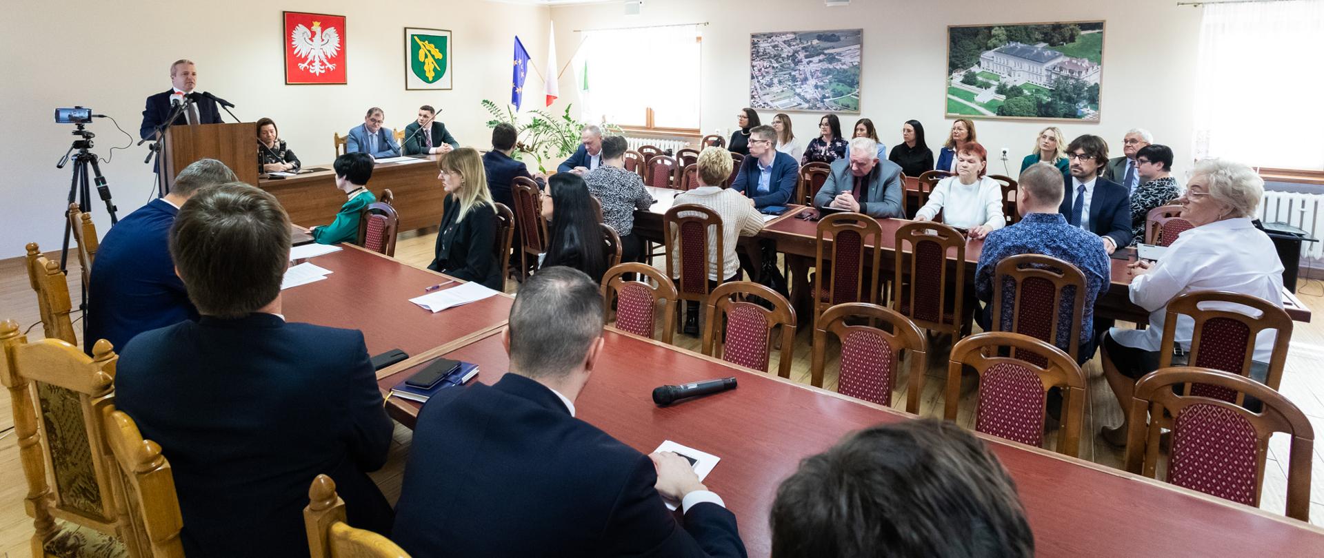 Jeden mężczyzna stojący przy mównicy i wypowiadający się. Przed nim wiele osób siedzących przy stołach