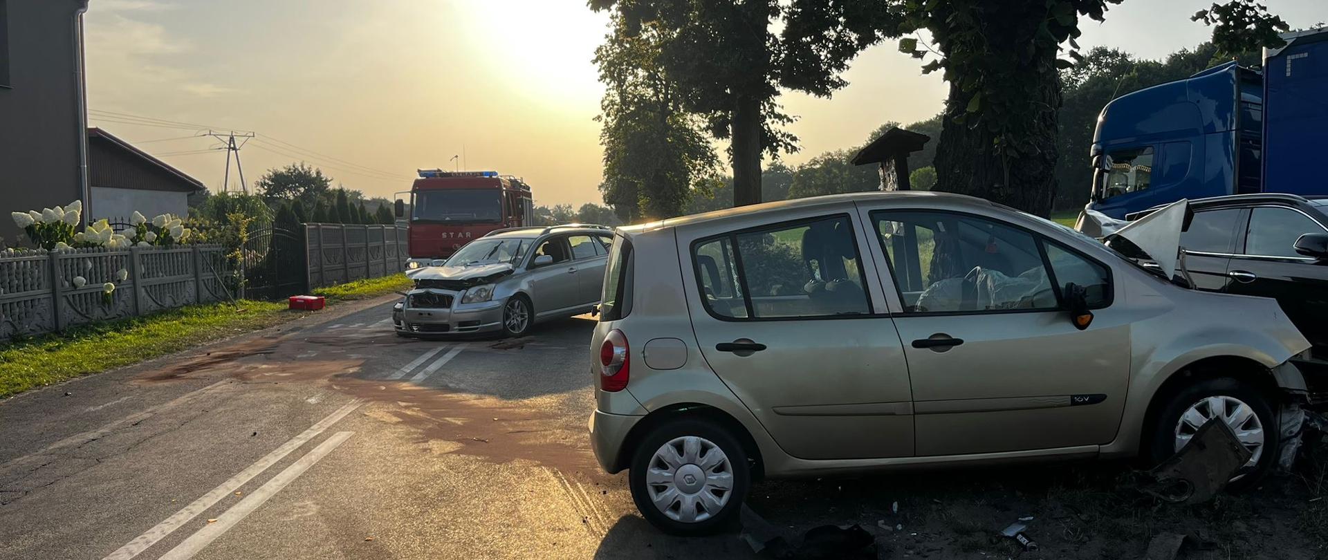 Złoty samochód osobowy po zderzeniu stoi przy drzewie na poboczu, obok niego drugi pojazd oraz jednostki straży pożarnej.