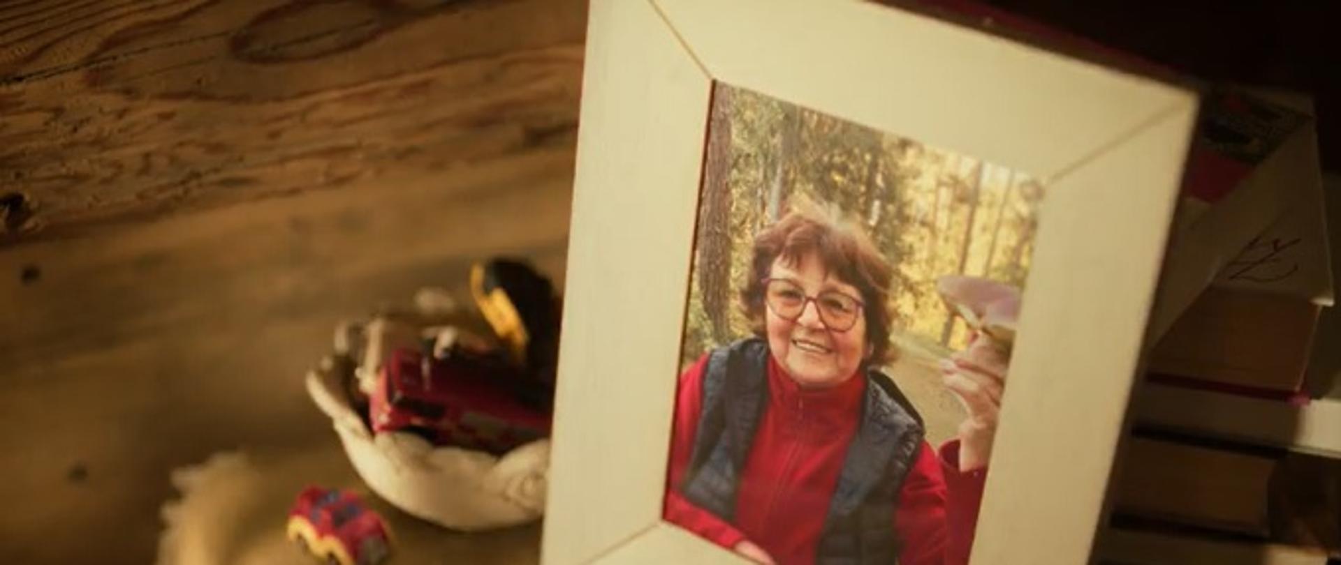 Zdjęcie przedstawia zdjęcie babci umieszczone na stoliku w ramce z podpisem babcia byłaby zadowolona. 