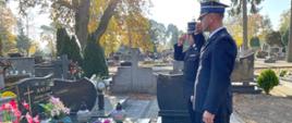 Dwóch strażaków w mundurach oddaje hołd przy grobie na cmentarzu